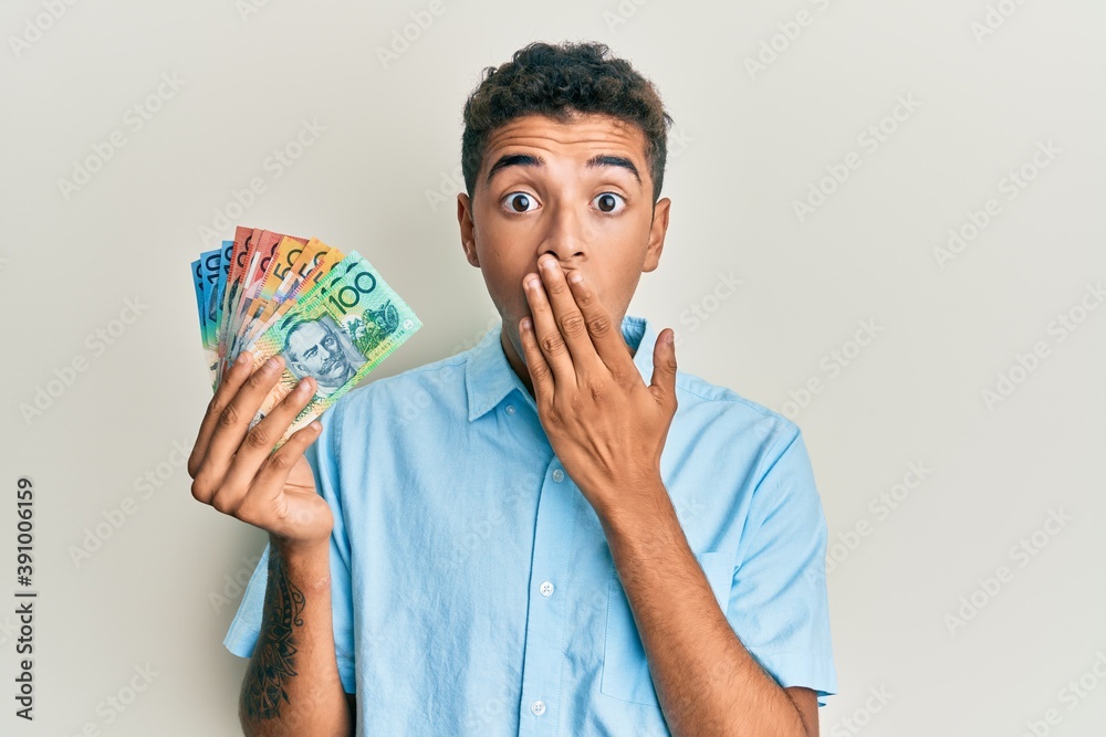Young handsome african american man holding australian dollars covering mouth with hand, shocked and afraid for mistake. surprised expression Young handsome african american man holding australian dollars covering mouth with hand, shocked and afraid for mistake. surprised expression