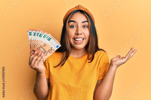 Young latin woman holding 50 euro banknotes celebrating achievement with happy smile and winner expression with raised hand