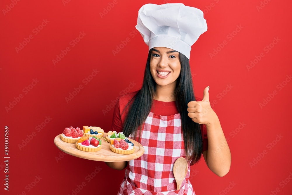 Young beautiful hispanic girl wearing baker apron holding cakes smiling happy and positive, thumb up doing excellent and approval sign Young beautiful hispanic girl wearing baker apron holding cakes smiling happy and positive, thumb up doing excellent and approval sign