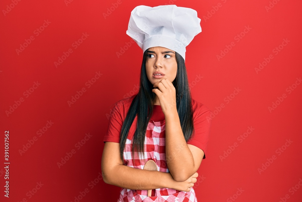 Young beautiful hispanic girl wearing baker uniform and cook hat serious face thinking about question with hand on chin, thoughtful about confusing idea Young beautiful hispanic girl wearing baker uniform and cook hat serious face thinking about question with hand on chin, thoughtful about confusing idea