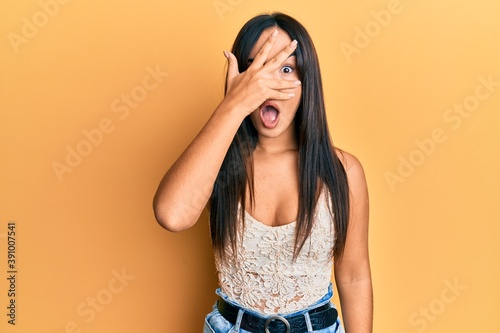Young beautiful hispanic girl wearing casual clothes peeking in shock covering face and eyes with hand, looking through fingers afraid