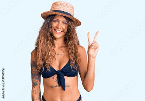 Young hispanic woman with tattoo wearing bikini and summer hat showing and pointing up with fingers number two while smiling confident and happy.