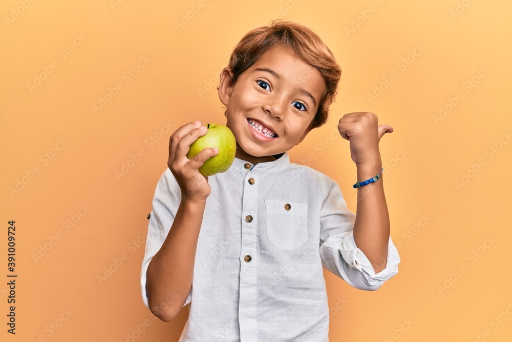 Adorable latin kid holding green apple pointing thumb up to the side smiling happy with open mouth - obrazy, fototapety, plakaty 