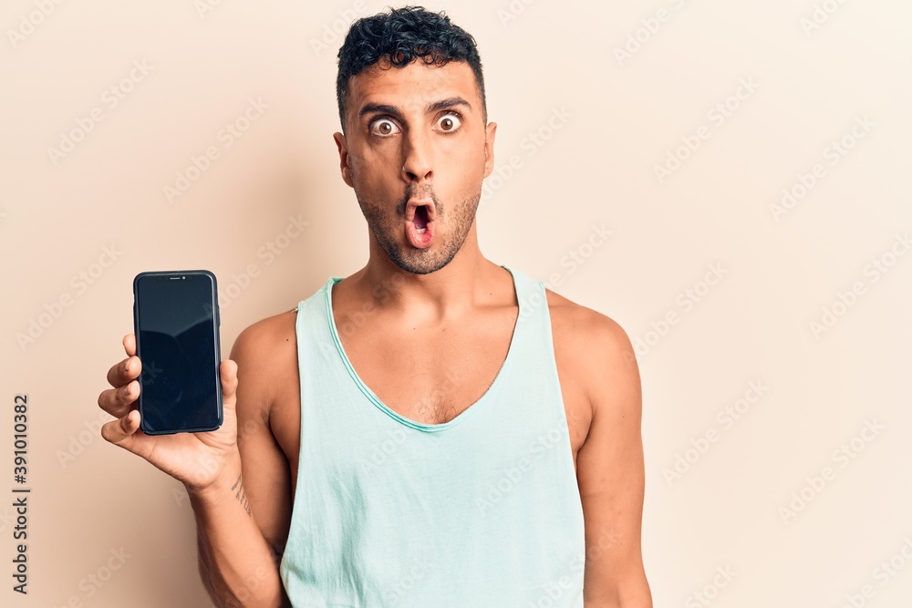 Young hispanic man holding smartphone showing screen scared and amazed with open mouth for surprise, disbelief face Young hispanic man holding smartphone showing screen scared and amazed with open mouth for surprise, disbelief face