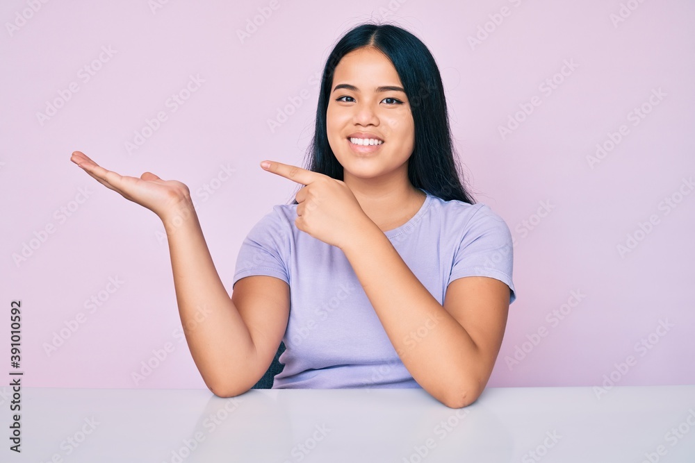 Young beautiful asian girl wearing casual clothes sitting on the table amazed and smiling to the camera while presenting with hand and pointing with finger. Young beautiful asian girl wearing casual clothes sitting on the table amazed and smiling to the camera while presenting with hand and pointing with finger.