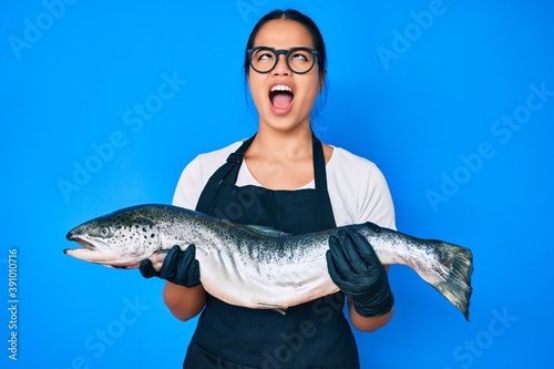 Young beautiful asian girl fishmonger selling fresh raw salmon angry and mad screaming frustrated and furious, shouting with anger looking up.