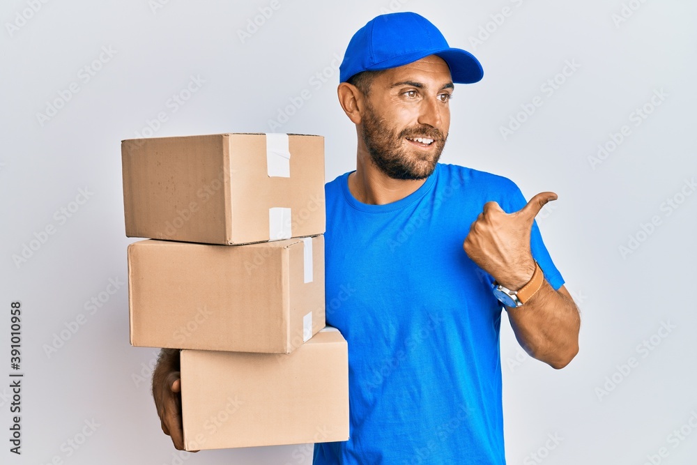 Handsome man with beard wearing courier uniform holding delivery packages smiling with happy face looking and pointing to the side with thumb up.