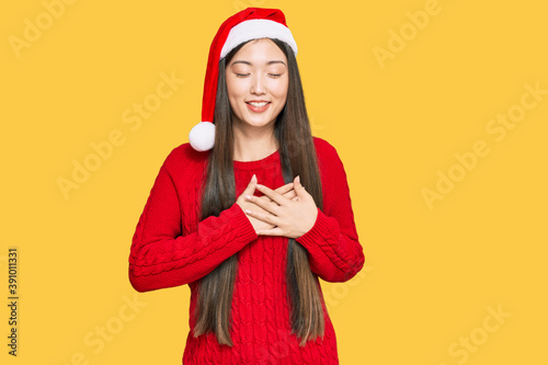 Young chinese woman wearing christmas hat smiling with hands on chest with closed eyes and grateful gesture on face. health concept.