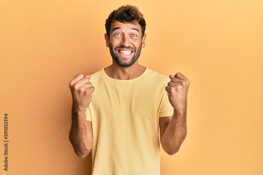 Handsome man with beard wearing casual yellow tshirt over yellow background celebrating surprised and amazed for success with arms raised and open eyes. winner concept.