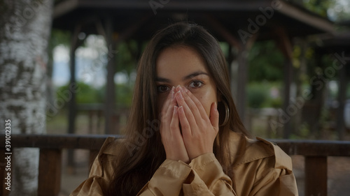 Charming pretty woman sitting in the gazebo, looks like she has a cold during flu season, looking at camera and sneezing and smiling, colds flu and covid-19 concept
