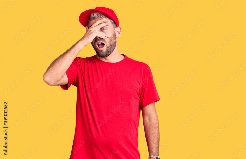 Fototapeta premium Young handsome blond man wearing t-shirt and cap peeking in shock covering face and eyes with hand, looking through fingers with embarrassed expression.