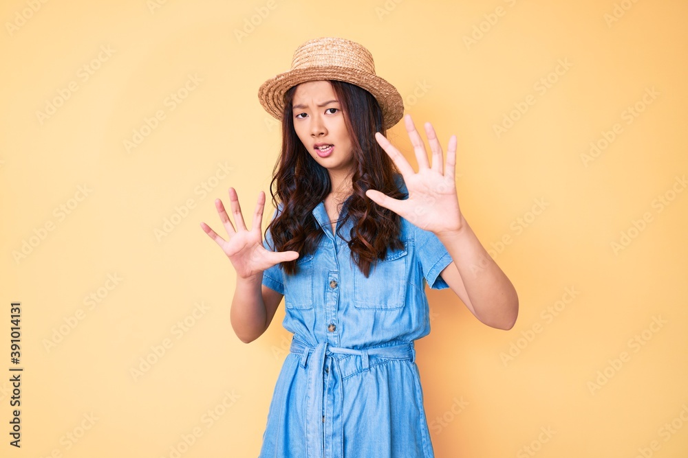 Young beautiful chinese girl wearing summer hat afraid and terrified with fear expression stop gesture with hands, shouting in shock. panic concept.