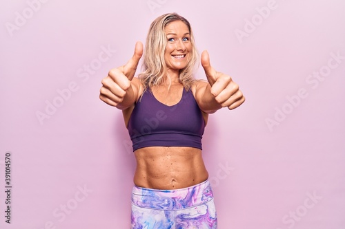 Middle age caucasian blonde woman wearing sportswear over pink background approving doing positive gesture with hand  thumbs up smiling and happy for success. winner gesture.