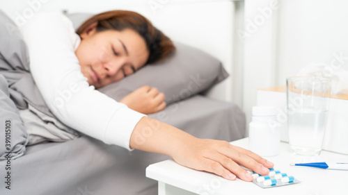sick Asian woman lying in bed take medicines pills and glass of water from bedside table to relieve symptoms.