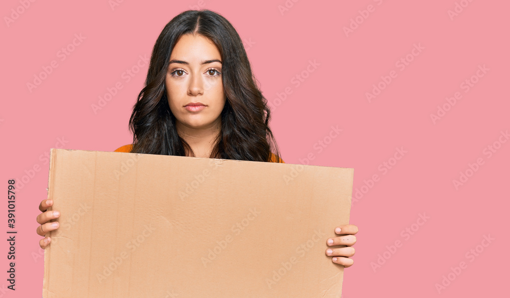 Beautiful brunette young woman holding cardboard blank empty banner thinking attitude and sober expression looking self confident Beautiful brunette young woman holding cardboard blank empty banner thinking attitude and sober expression looking self confident
