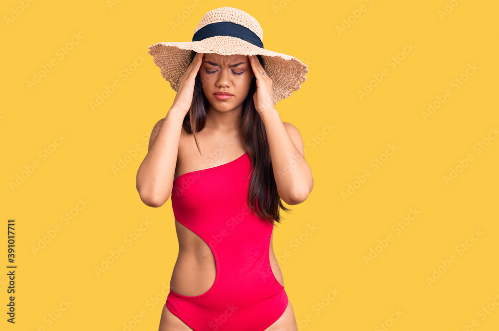 Young beautiful latin girl wearing swimwear and summer hat with hand on head for pain in head because stress. suffering migraine. Young beautiful latin girl wearing swimwear and summer hat with hand on head for pain in head because stress. suffering migraine.