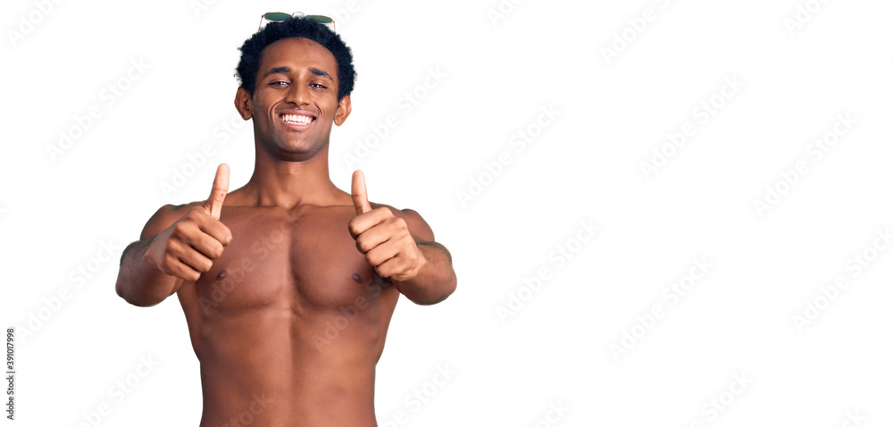 African handsome man wearing swimsuit and sunglasses approving doing positive gesture with hand, thumbs up smiling and happy for success. winner gesture.