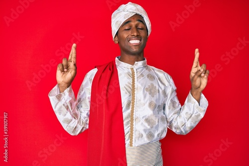 Handsome indian man wearing tradition sherwani saree clothes gesturing finger crossed smiling with hope and eyes closed. luck and superstitious concept.