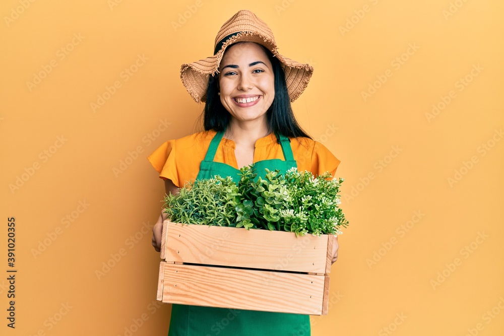 Beautiful young woman wearing gardener apron holding wooden plant pot smiling with a happy and cool smile on face. showing teeth.