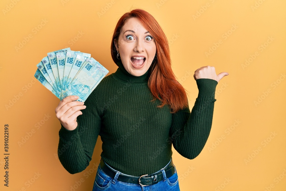 Beautiful redhead woman holding 100 brazilian real banknotes pointing thumb up to the side smiling happy with open mouth Beautiful redhead woman holding 100 brazilian real banknotes pointing thumb up to the side smiling happy with open mouth