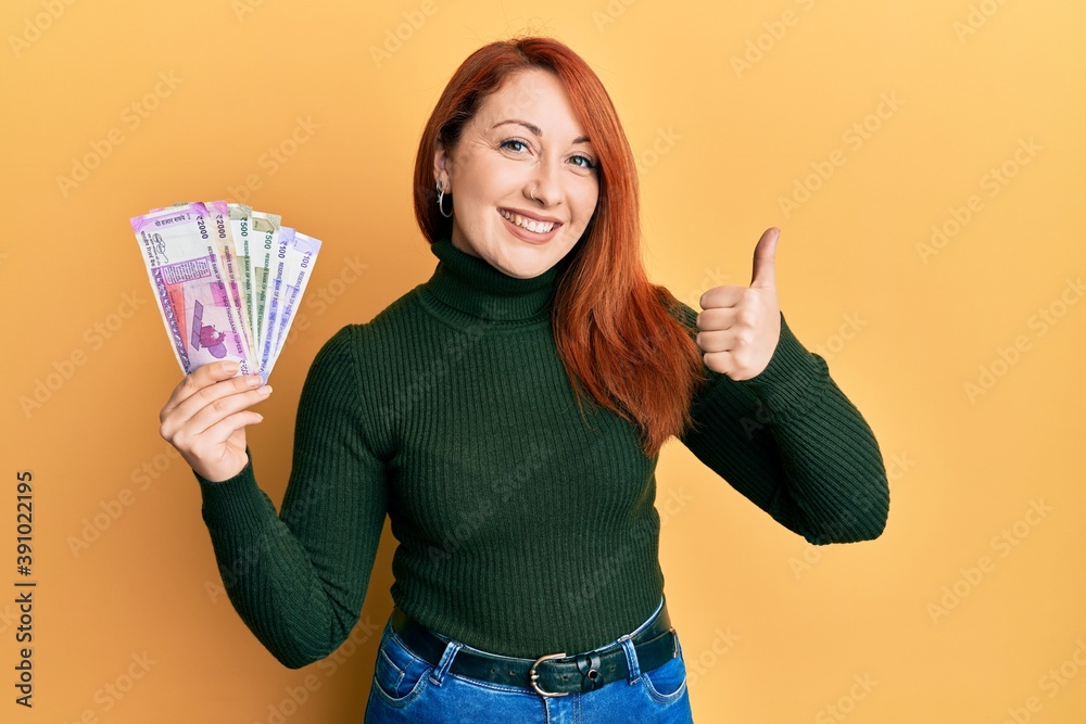Beautiful redhead woman holding indian rupee banknotes smiling happy and positive, thumb up doing excellent and approval sign Beautiful redhead woman holding indian rupee banknotes smiling happy and positive, thumb up doing excellent and approval sign