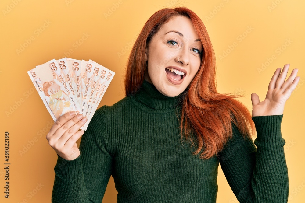 Beautiful redhead woman holding 50 turkish lira banknotes celebrating achievement with happy smile and winner expression with raised hand Beautiful redhead woman holding 50 turkish lira banknotes celebrating achievement with happy smile and winner expression with raised hand