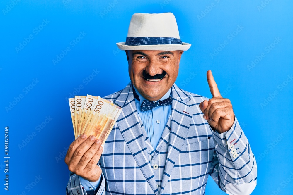 Mature middle east man with mustache wearing elegant vintage style holding norwegian krone smiling with an idea or question pointing finger with happy face, number one Mature middle east man with mustache wearing elegant vintage style holding norwegian krone smiling with an idea or question pointing finger with happy face, number one