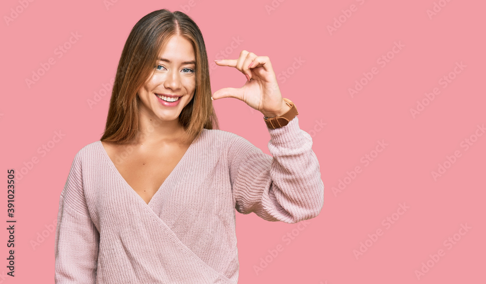 Beautiful blonde woman wearing casual winter pink sweater smiling and confident gesturing with hand doing small size sign with fingers looking and the camera. measure concept.