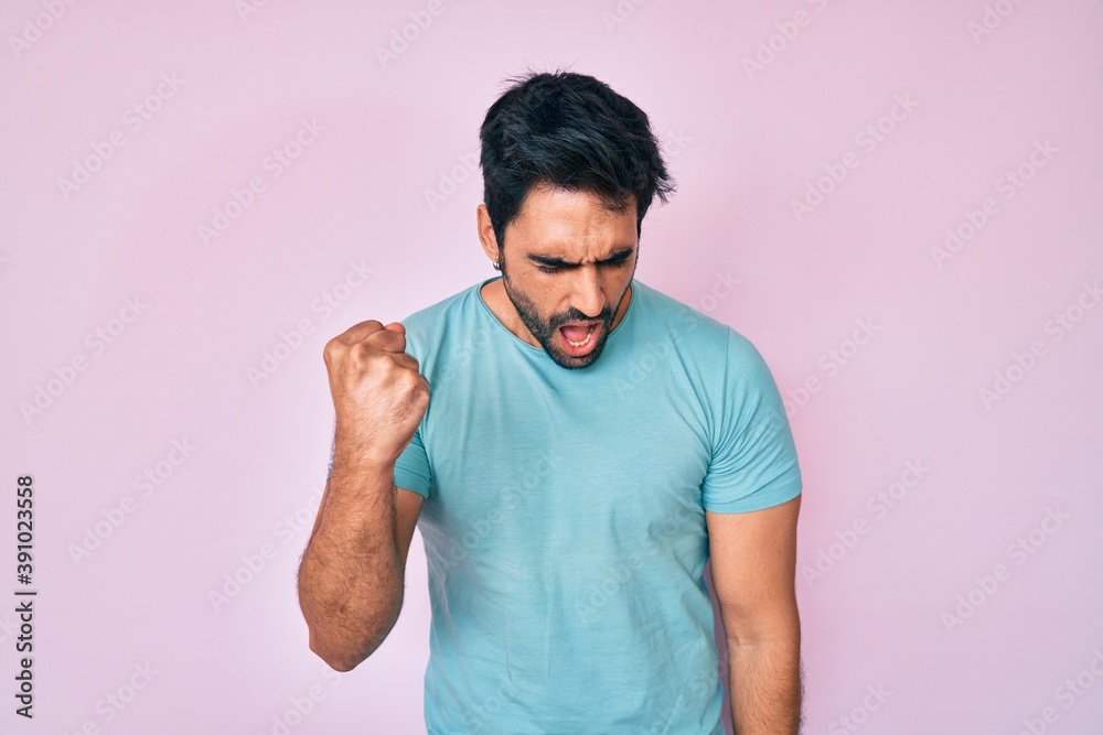 Handsome hispanic man with beard wearing casual clothes angry and mad raising fist frustrated and furious while shouting with anger. rage and aggressive concept. Handsome hispanic man with beard wearing casual clothes angry and mad raising fist frustrated and furious while shouting with anger. rage and aggressive concept.