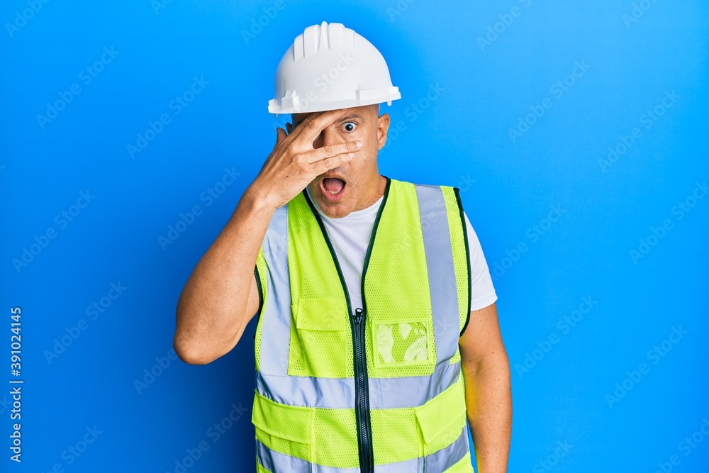 Middle age bald man wearing architect hardhat peeking in shock covering face and eyes with hand, looking through fingers afraid