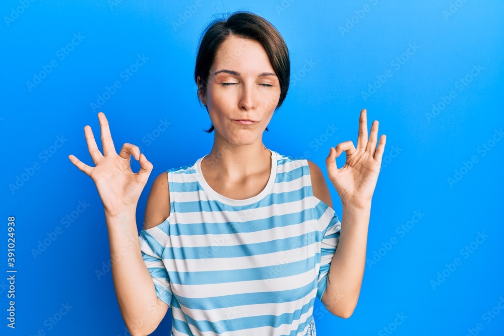 Young brunette woman with short hair wearing casual striped t-shirt relax and smiling with eyes closed doing meditation gesture with fingers. yoga concept. Young brunette woman with short hair wearing casual striped t-shirt relax and smiling with eyes closed doing meditation gesture with fingers. yoga concept.