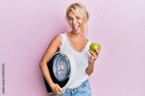 Young blonde girl holding weight machine and green apple sticking tongue out happy with funny expression.