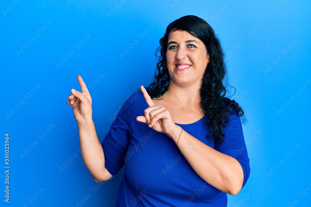 Fototapeta premium Plus size brunette woman wearing casual blue shirt smiling and looking at the camera pointing with two hands and fingers to the side.