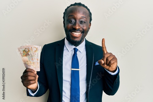 Handsome young black man wearing business suit holding pounds banknotes smiling with an idea or question pointing finger with happy face, number one