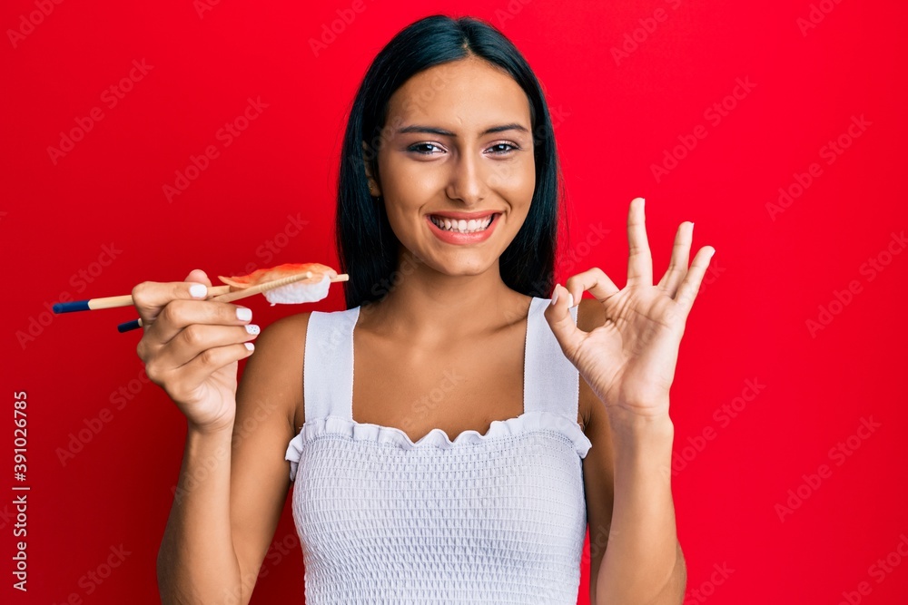 Young brunette woman eating prawn sushi using chopsticks doing ok sign with fingers, smiling friendly gesturing excellent symbol