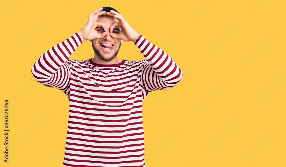 Young handsome man wearing striped sweater doing ok gesture like binoculars sticking tongue out, eyes looking through fingers. crazy expression. Young handsome man wearing striped sweater doing ok gesture like binoculars sticking tongue out, eyes looking through fingers. crazy expression.