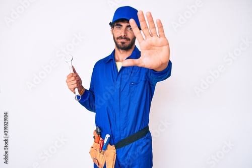 Handsome young man with curly hair and bear wearing builder jumpsuit uniform and holding wrench with open hand doing stop sign with serious and confident expression, defense gesture