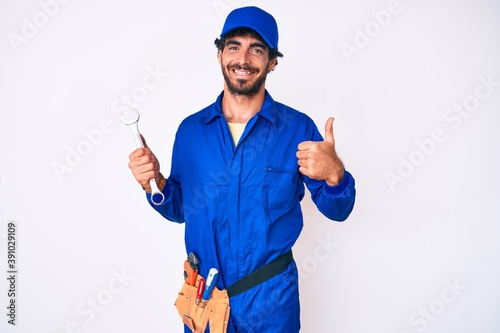 Handsome young man with curly hair and bear wearing builder jumpsuit uniform and holding wrench smiling happy and positive, thumb up doing excellent and approval sign