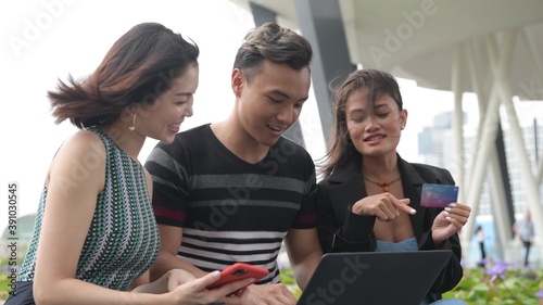 Three asian friends seated on a city bench making shopping on the web, buying from laptop © jovannig