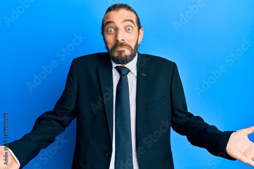 Attractive man with long hair and beard wearing business suit and tie clueless and confused expression with arms and hands raised. doubt concept.