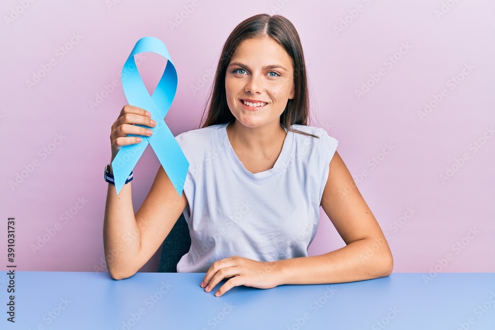 Young caucasian woman holding blue ribbon looking positive and happy standing and smiling with a confident smile showing teeth Young caucasian woman holding blue ribbon looking positive and happy standing and smiling with a confident smile showing teeth