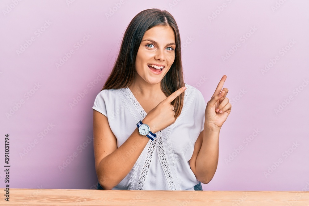 Young caucasian woman wearing casual clothes sitting on the table smiling and looking at the camera pointing with two hands and fingers to the side. Young caucasian woman wearing casual clothes sitting on the table smiling and looking at the camera pointing with two hands and fingers to the side.