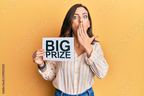 Young brunette woman holding big prize paper covering mouth with hand, shocked and afraid for mistake. surprised expression