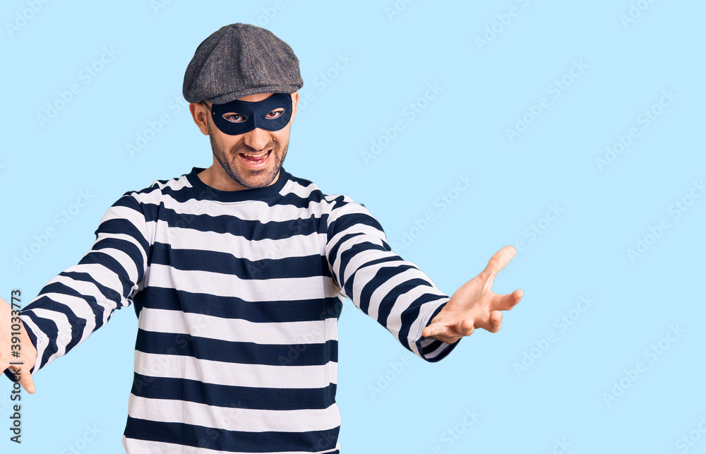 Young handsome man wearing burglar mask looking at the camera smiling with open arms for hug. cheerful expression embracing happiness. Young handsome man wearing burglar mask looking at the camera smiling with open arms for hug. cheerful expression embracing happiness.