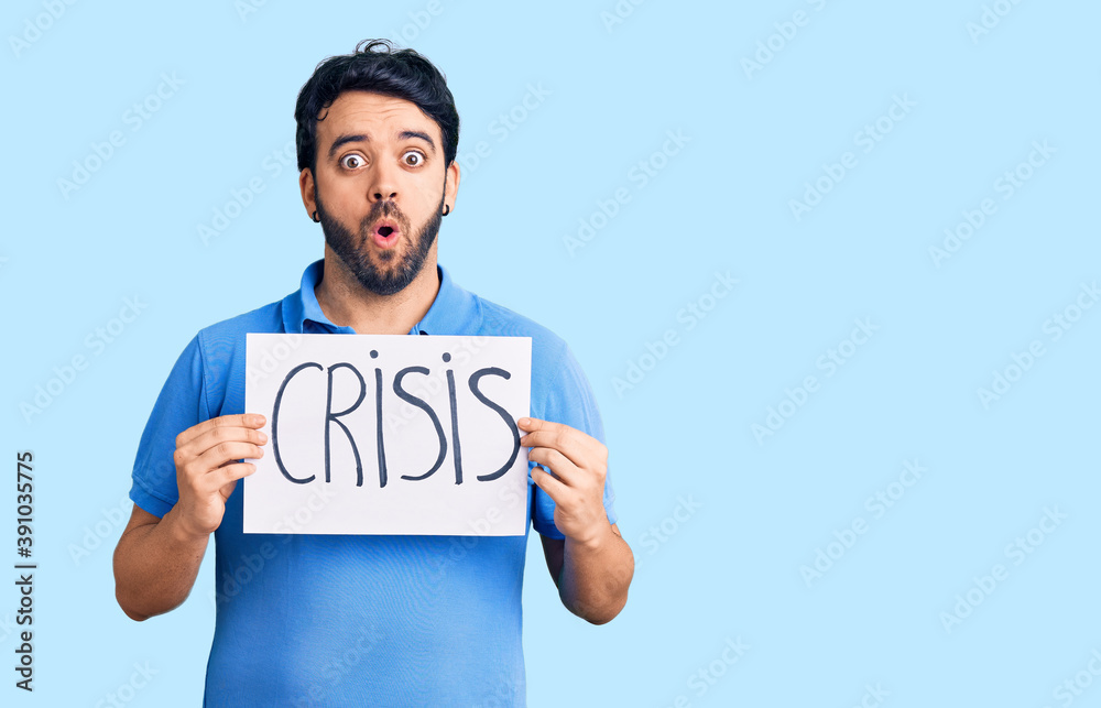 Young hispanic man holding crisis message paper scared and amazed with open mouth for surprise, disbelief face Young hispanic man holding crisis message paper scared and amazed with open mouth for surprise, disbelief face