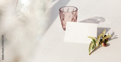 Summer stationery still life scene.Glass of water in sunlight with harsh shadows on white background, olive branch. Blank greeting card, invitation mockup scene Flat lay,top view.Modern minimal design