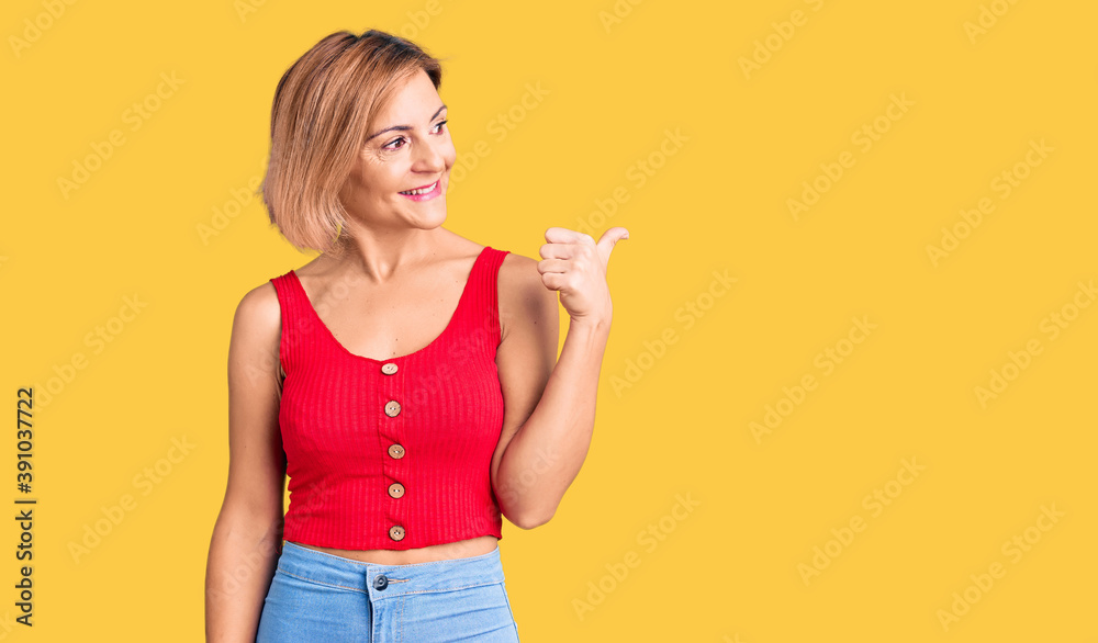 Young blonde woman wearing casual clothes smiling with happy face looking and pointing to the side with thumb up. Young blonde woman wearing casual clothes smiling with happy face looking and pointing to the side with thumb up.