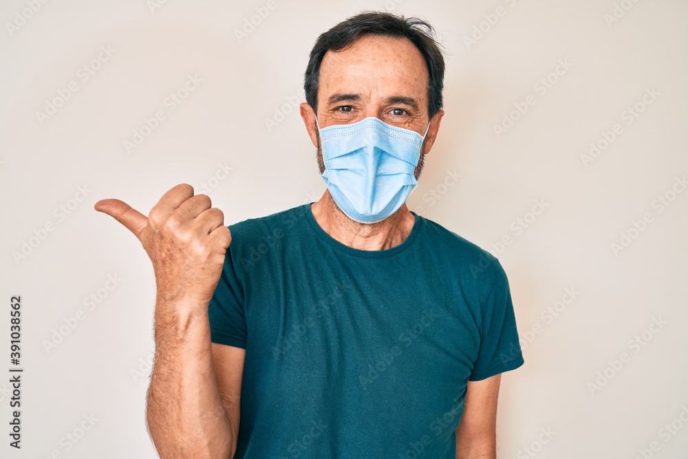 Middle age hispanic man wearing medical mask pointing thumb up to the side smiling happy with open mouth Middle age hispanic man wearing medical mask pointing thumb up to the side smiling happy with open mouth