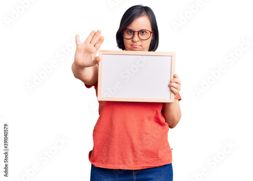 Brunette woman with down syndrome holding empty white chalkboard with open hand doing stop sign with serious and confident expression, defense gesture