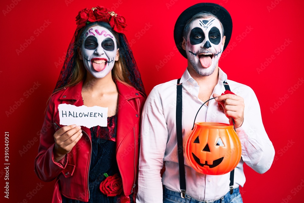 Couple wearing day of the dead costume holding pumpking and halloween paper sticking tongue out happy with funny expression.